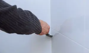 Ręka w swetrze usuwa stary silikon z narożnika białych płytek za pomocą narzędzia.