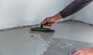 Ręka z kielnią rozprowadza masę samopoziomującą. Trudno ocenić, jaka grubość wylewki samopoziomującej jest tu stosowana.
