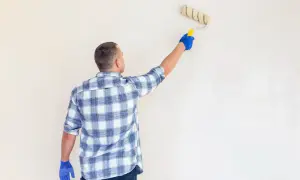 Mężczyzna w kratę nakłada grunt pod tynki gipsowe na ścianę wałkiem.
