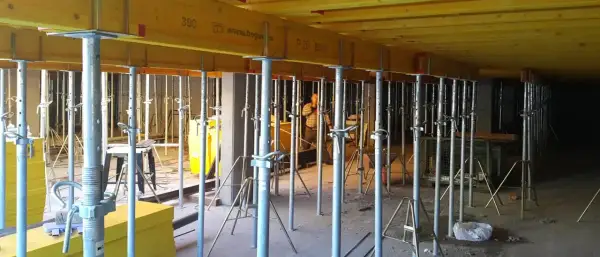 Metalowe stemple budowlane podtrzymują żółte belki stropowe. W tle widać pracownika i inne elementy konstrukcyjne.