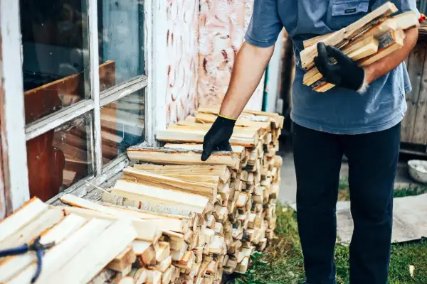 Mężczyzna w rękawiczkach układa drewno przy oknie. Zastanawia się, ile kosztuje kubik drzewa na zimę.
