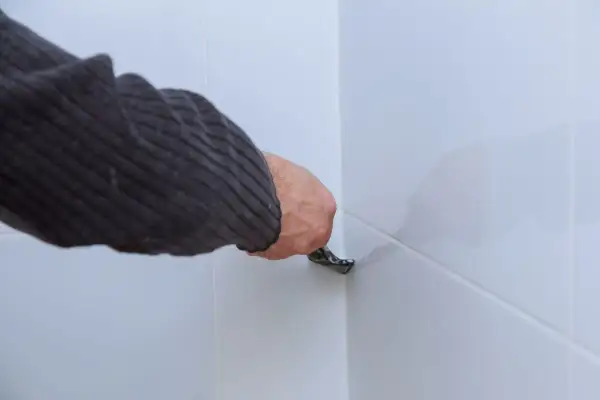 Ręka w swetrze usuwa stary silikon z narożnika białych płytek za pomocą narzędzia.